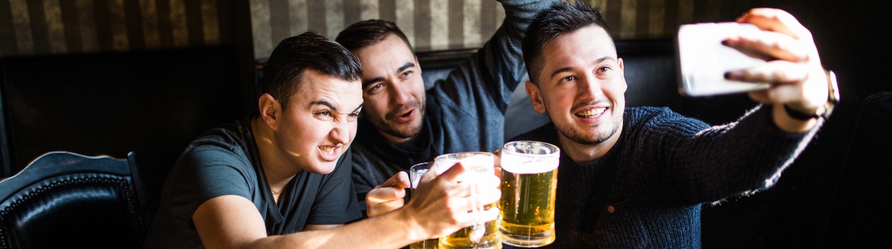 A group of men toasting beers and one of them is taking a group selfie